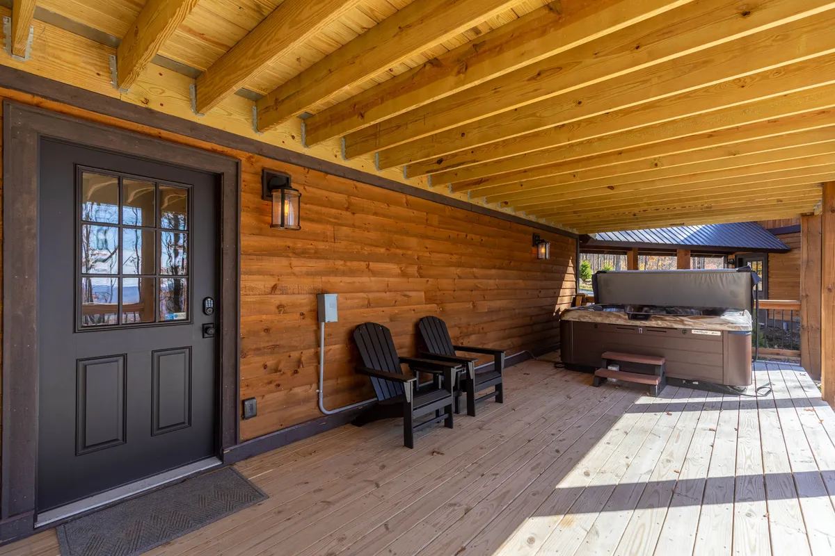 The image shows the exterior of a luxury mountain home featuring a welcoming front porch with wooden chairs and a private hot tub. The tranquil setting is surrounded by natural landscape, evoking relaxation and a connection to the outdoors.