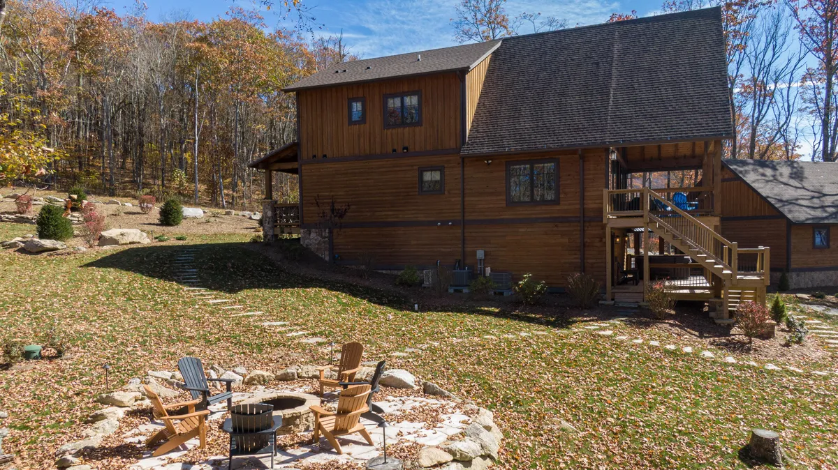 A cozy fire pit area with Adirondack chairs encircles a stone fire pit, set beside the wooden house and bordered by a stone pathway and surrounding trees. The inviting space is ideal for storytelling, s’mores, and starlit gatherings.