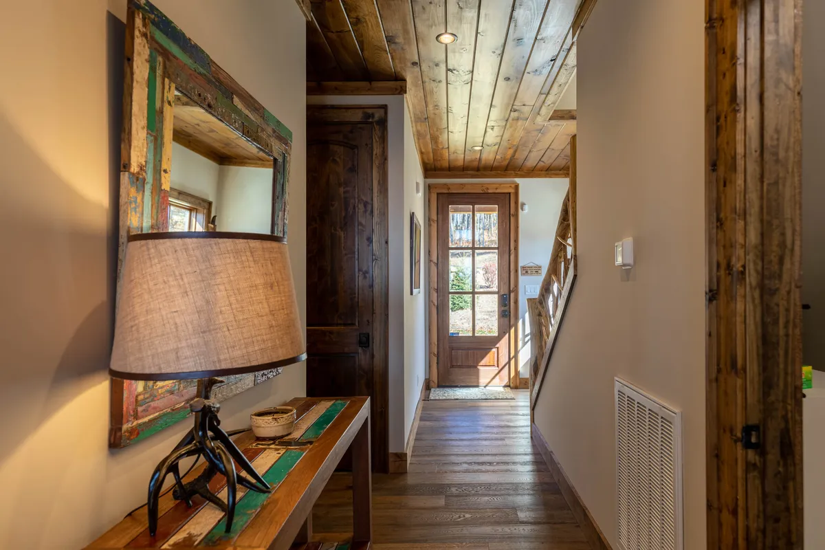 This image features the wooden front door of the property, a decorative mirror, and a table lamp. The entryway is warm and inviting, setting the tone for a welcoming arrival. Natural light hints at the outdoors, and the space feels ready for guests and their four-legged companions to come in from a mountain adventure.