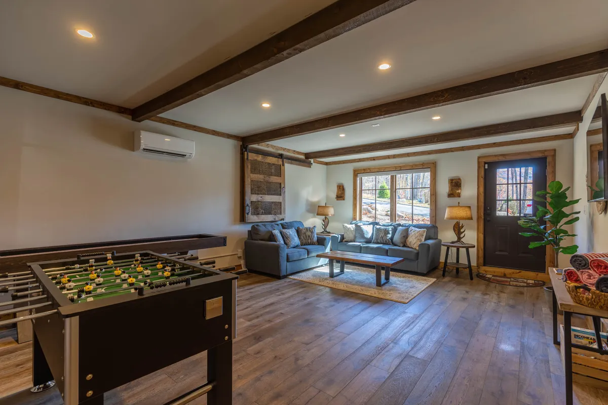 Sunlight streams through a large window into the game room, which showcases a foosball table and a comfortable sofa. The wooden door and ample natural light create a welcoming, casual atmosphere ideal for both active play and relaxation.