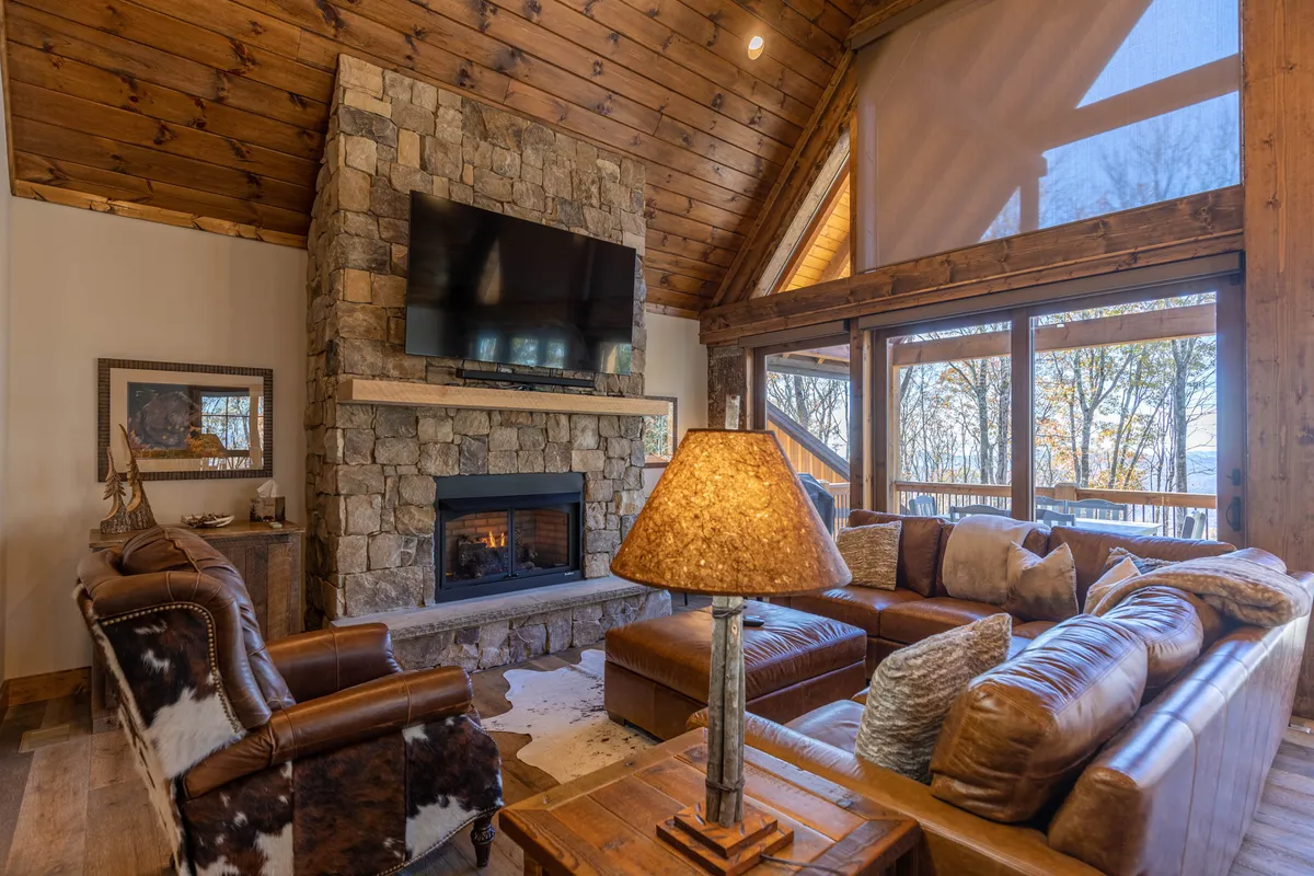 This spacious living area showcases leather sofas gathered around a stone fireplace, with large windows framing beautiful mountain views. Wooden beams and a decorative lamp add rustic charm, while the arrangement suggests a cozy spot for guests and pets to relax after a day outdoors.