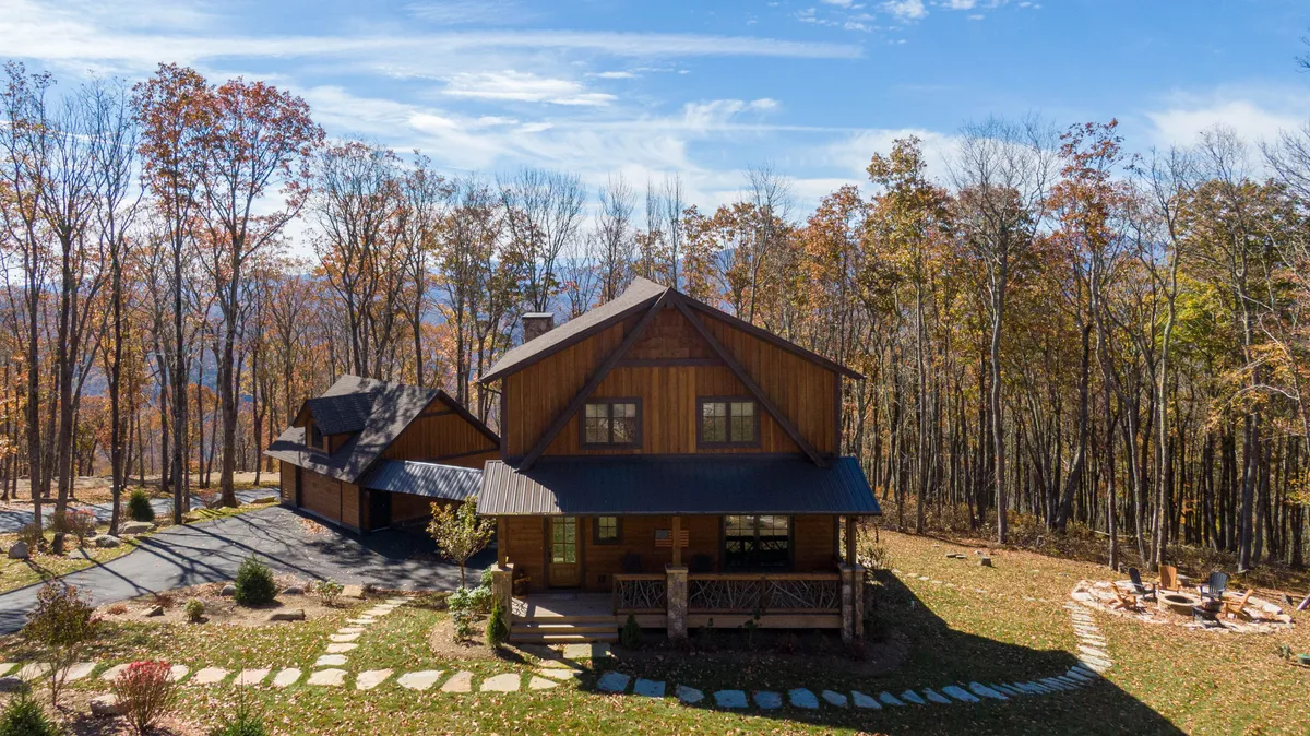 An inviting exterior image with a wooden house nestled among trees, a cozy fire pit area with seating, and a stone pathway weaving through the yard. The lush surroundings and comfortable seating suggest a perfect spot for evening gatherings, s’mores, and stargazing in a mountain setting.