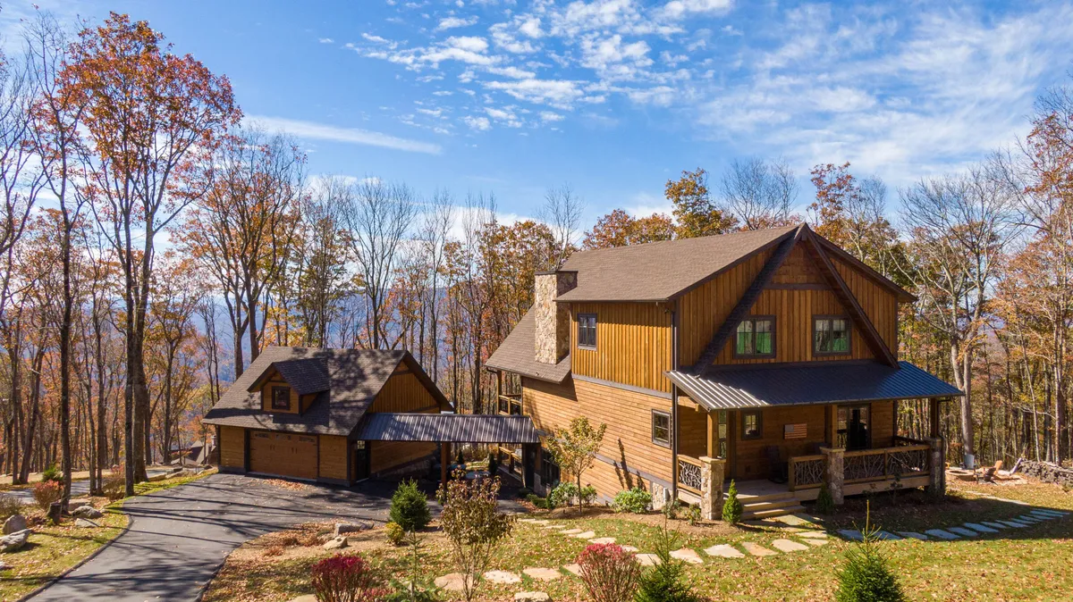 The image showcases the wooden cabin exterior nestled among tall trees, with a welcoming stone pathway leading up to the front and an attached garage. The porch offers scenic mountain sunrise views, and the tranquil setting hints at opportunities for bonfires and wildlife observation.
