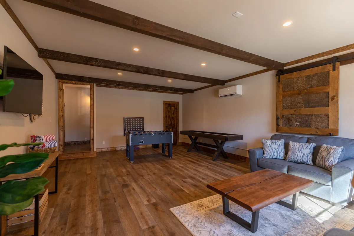 A versatile lounge space with a foosball table, comfortable sofa, TV, and wooden beams overhead. The room is designed for both relaxation and activity, capturing the dual purpose of the lower level as a hub for game nights and cozy movie evenings.