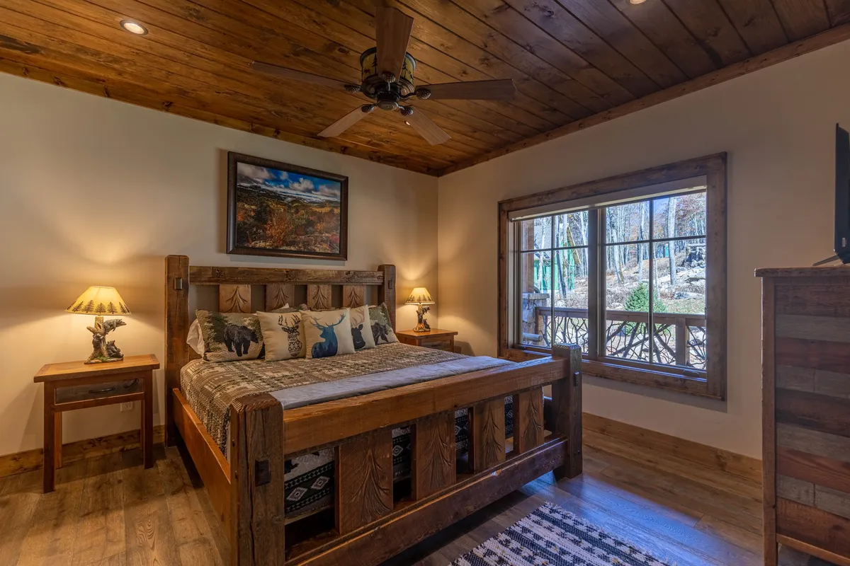 A tranquil bedroom featuring a wooden bed frame with plush pillows, tasteful artwork on the wall, and a window that lets in natural light and offers a view of the outdoors. The room exudes calmness and comfort, perfect for restful nights.