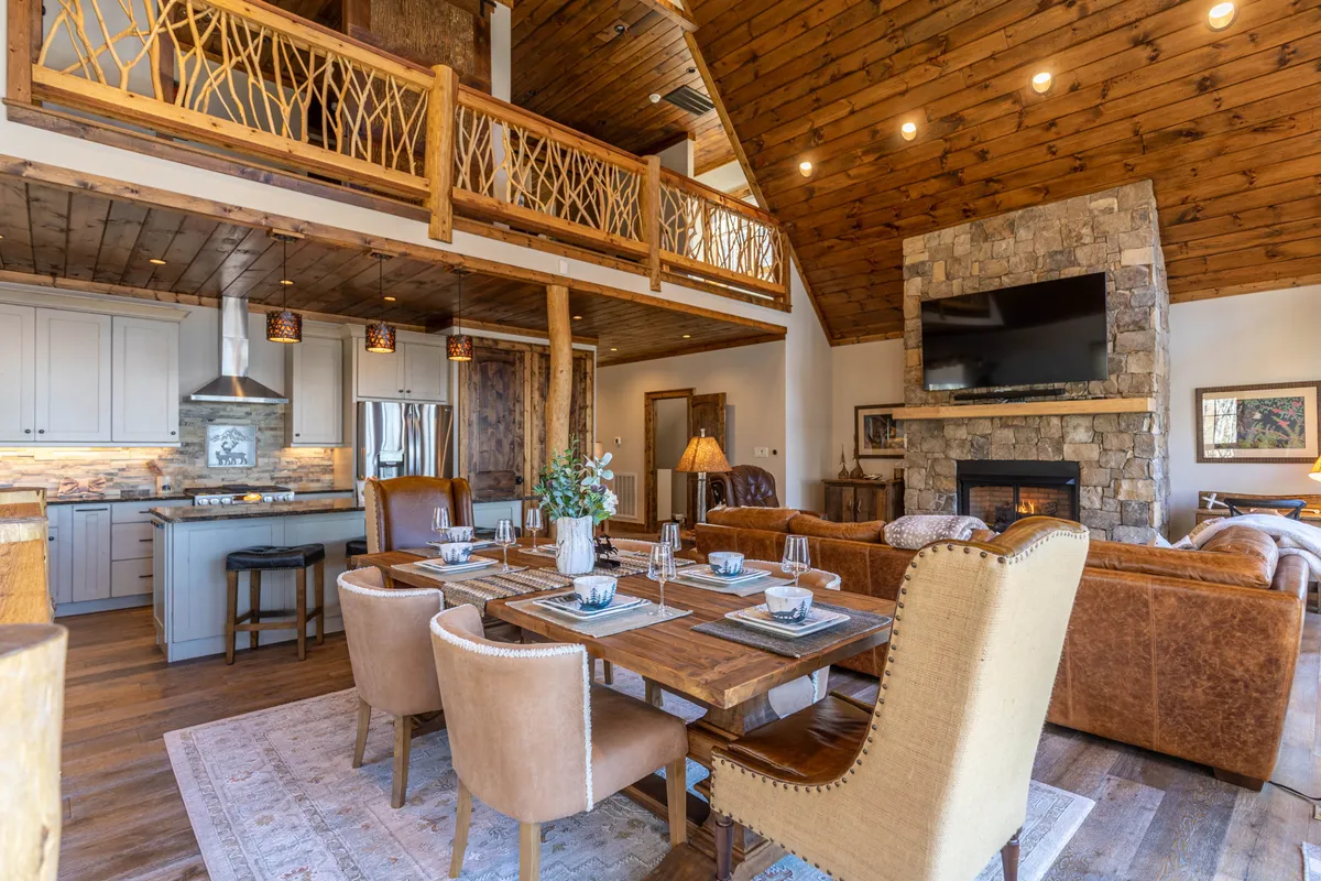 An open-plan dining and living area anchored by a large wooden dining table, a stone fireplace, visible kitchen space, and broad wooden beams. The cozy setting is ideal for shared meals, special gatherings, and relaxing by the fire, with a seamless flow between spaces.