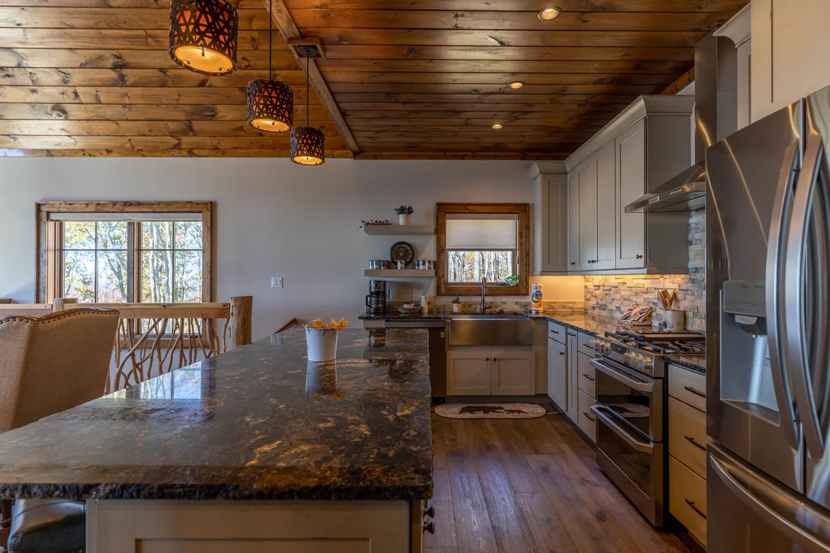 A bright, modern kitchen featuring expansive granite countertops, warm wooden beams overhead, pendant lighting, a full suite of stainless steel appliances, and a window offering a mountain view. The space is both functional and stylish, with custom cabinetry and a layout perfect for cooking and socializing.