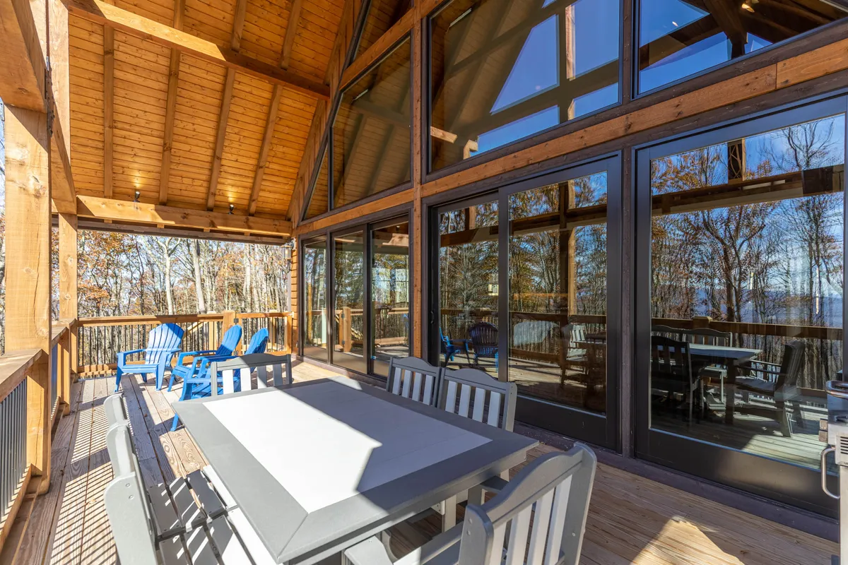 The image shows a spacious wooden deck with a large outdoor dining table and blue Adirondack chairs, set against a backdrop of large glass windows. The deck overlooks serene natural surroundings, inviting guests to relax, enjoy meals outdoors, and take in the fresh mountain air.